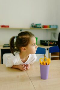 A young girl in a classroom setting, focused and attentive, with scissors on the table.